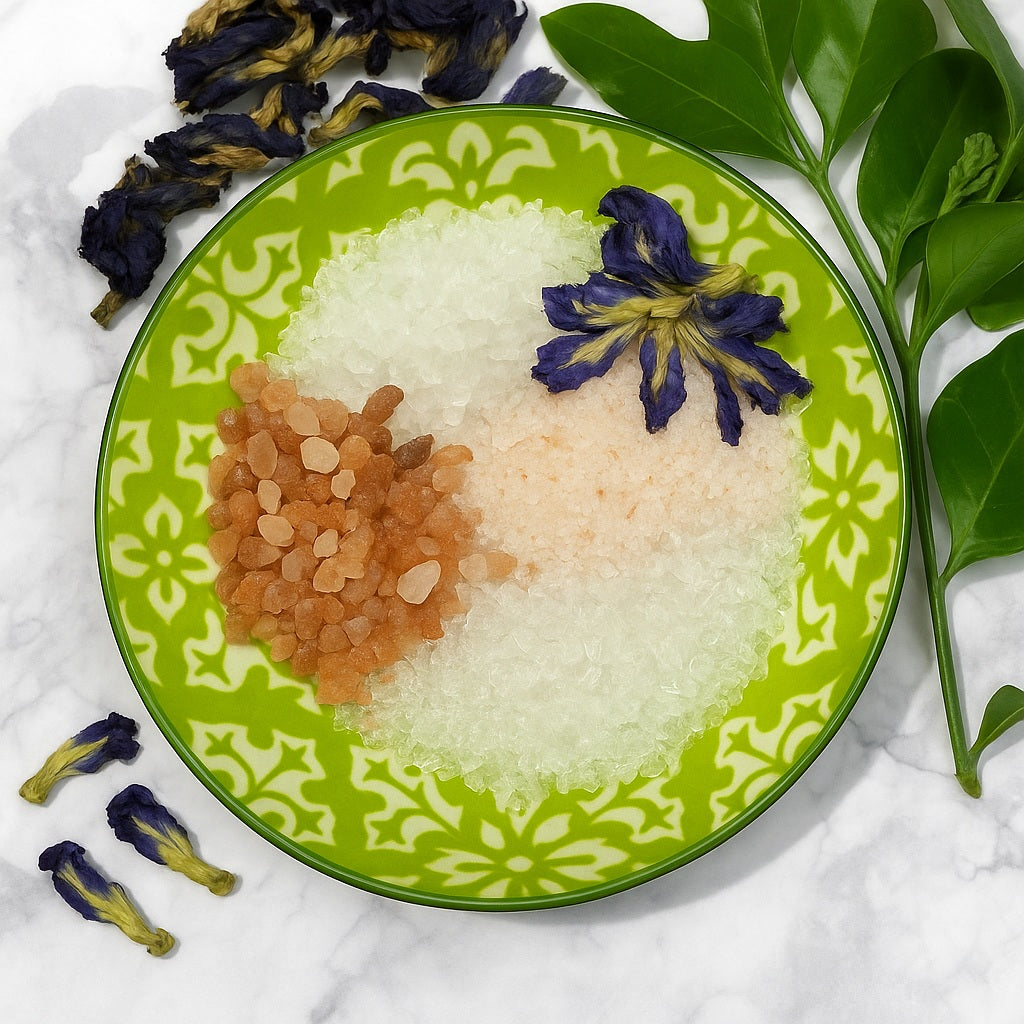 A green and white patterned ceramic bowl filled with bath salts and topped with dried butterfly pea flowers, set on a marble surface with green leaves and scattered dried flowers around it.