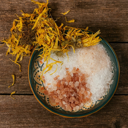 A ceramic bowl filled with coarse white, pink, and Himalayan salts, topped with dried calendula flowers, set on a rustic wooden bench background.