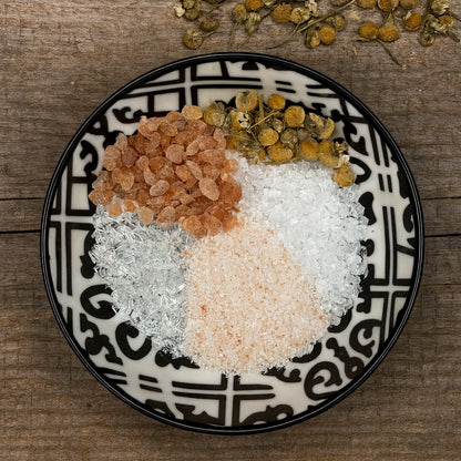 A black and white patterned ceramic bowl filled with various salts and dried chamomile flowers, placed on a rustic wooden bench background.