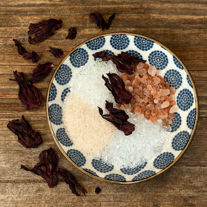 A blue and white patterned ceramic bowl filled with assorted bath salts and dried hibiscus flowers, set on a rustic wooden bench with scattered hibiscus petals around it.