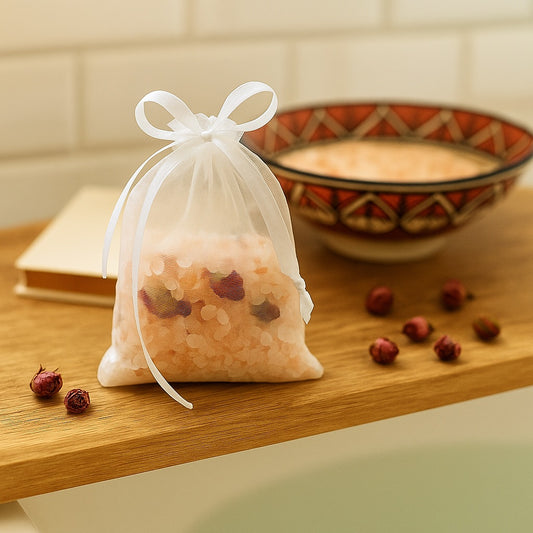 Transparent organza bag filled with pink Himalayan salt and rose buds, tied with a bow, placed on a wooden bath tray.