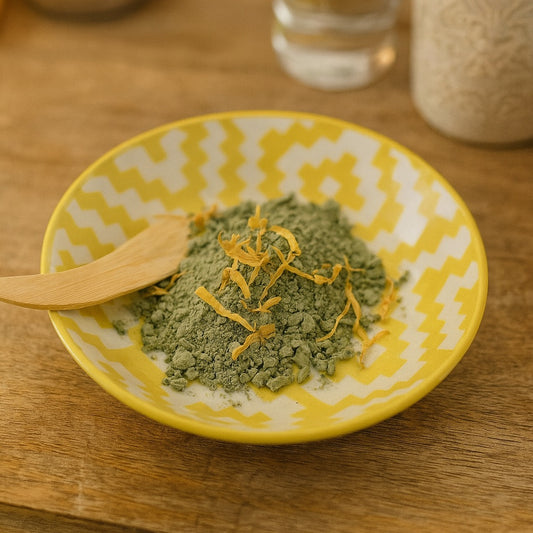 A yellow patterned ceramic bowl filled with fine French green clay powder, accented with scattered dried calendula petals, placed on a rustic wooden vanity with warm lighting and subtle country chic decor in the background.