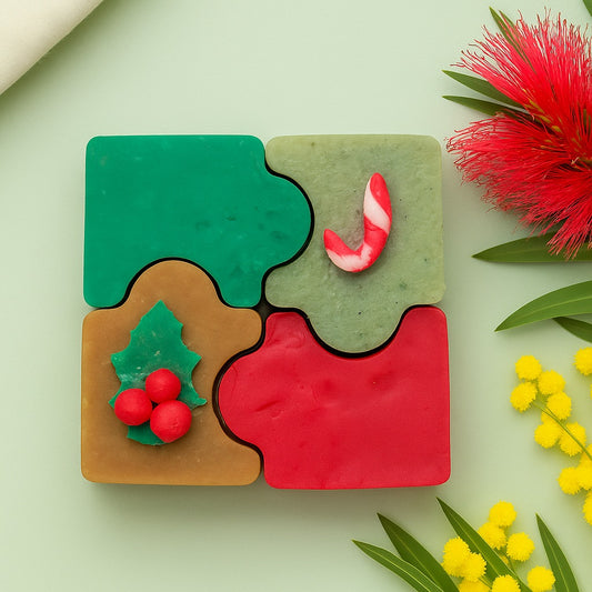 Four Christmas-themed puzzle soaps arranged together on a festive background with red bottlebrush and yellow wattle flowers, bright natural lighting, country chic style.
