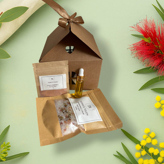 Little Luxe Stocking Trio from Terra’s Touch displayed on a soft green botanical background with wattle and bottlebrush. Includes a Clay & Chamomile dry face mask, a small bottle of golden facial oil, and a Chamomile bath soak presented beside a kraft house-shaped gift box tied with a satin ribbon.