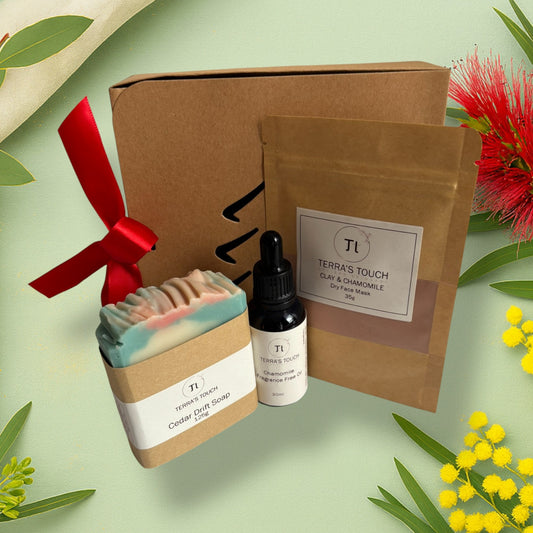 Gentle Ritual Set from Terra’s Touch displayed on a soft green botanical background with wattle and bottlebrush. Includes a Cedar Drift soap bar, Chamomile fragrance-free face oil, and a Clay & Chamomile dry face mask presented beside a kraft gift box with a Christmas tree cut-out and red ribbon bow.