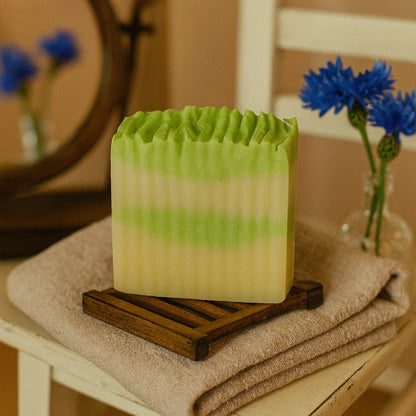 Handmade green clay soap bar with textured top, placed on a wooden soap saver in a country chic setting with rustic décor accents