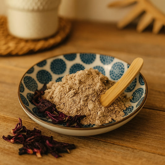 A ceramic dish filled with Moroccan clay powder and a wooden spatula, accented with dried hibiscus flowers, set on a rustic wooden vanity with warm lighting and subtle country chic décor in the background.