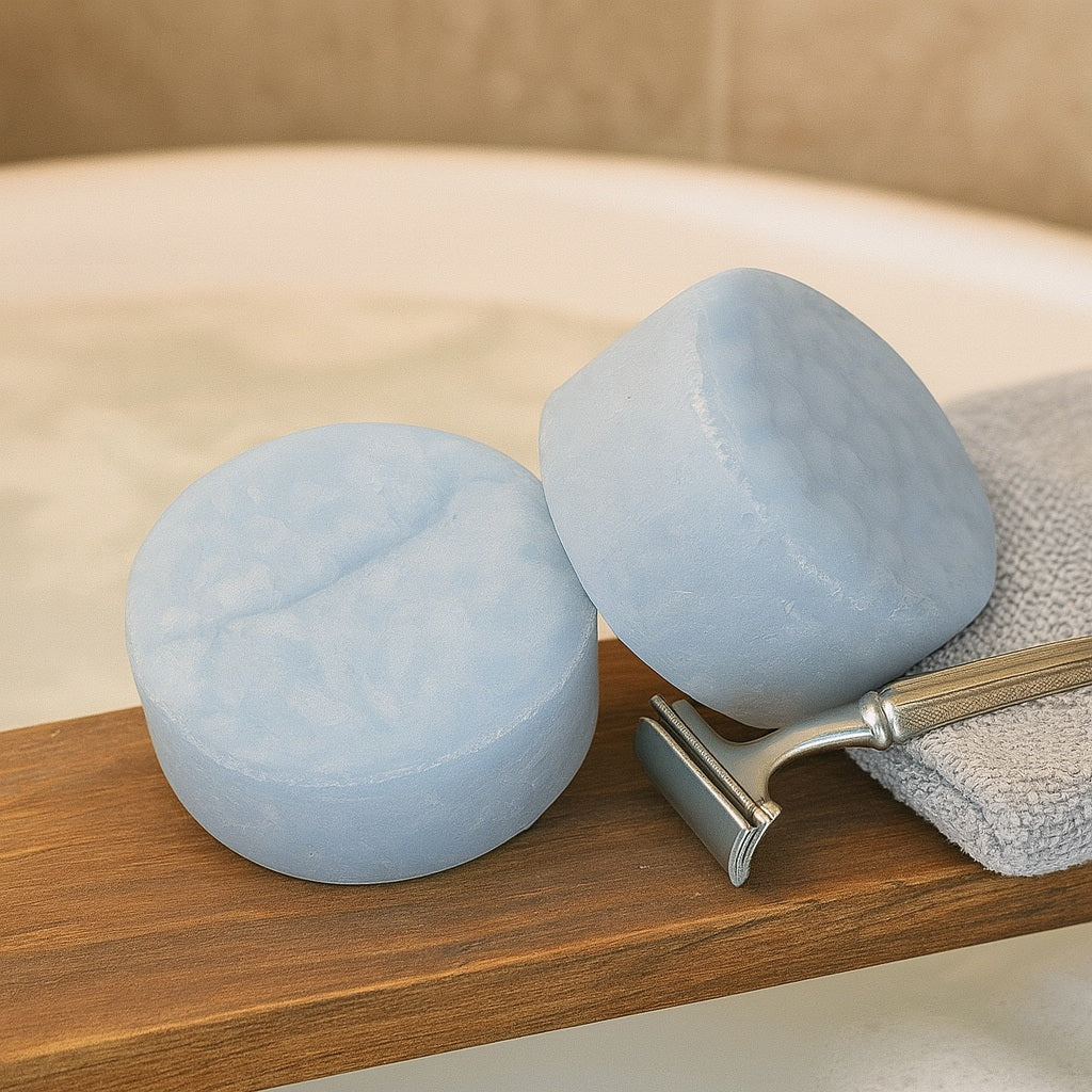 wo pale blue-grey Sandalwood Nut Shampoo Bars from Terra’s Touch displayed on a rustic wooden bath shelf above a frothy milky tub, styled with a folded towel and a classic razor for a subtle masculine country chic look.