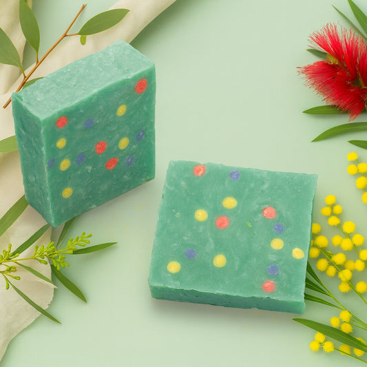 Two handmade mint-green soap bars with colorful confetti dots, displayed on a soft Christmas background featuring native Australian flora and festive greenery, styled in a warm country-chic theme.