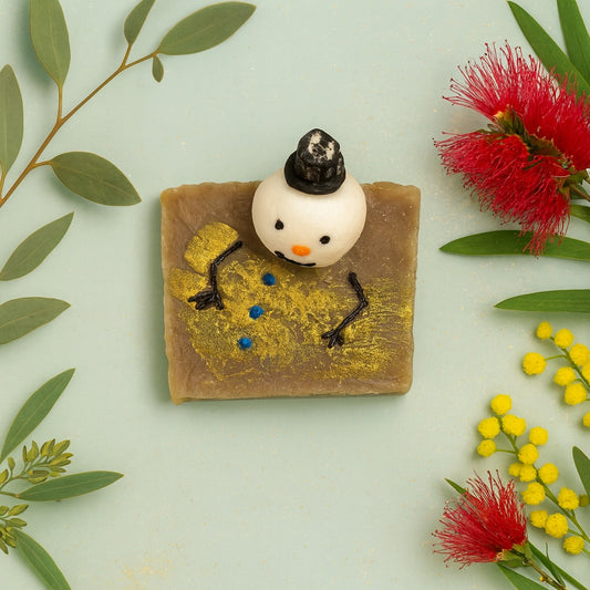 Handmade artisan soap bar featuring a small snowman figure on top, with golden shimmer detailing and a neutral background resembling the beach