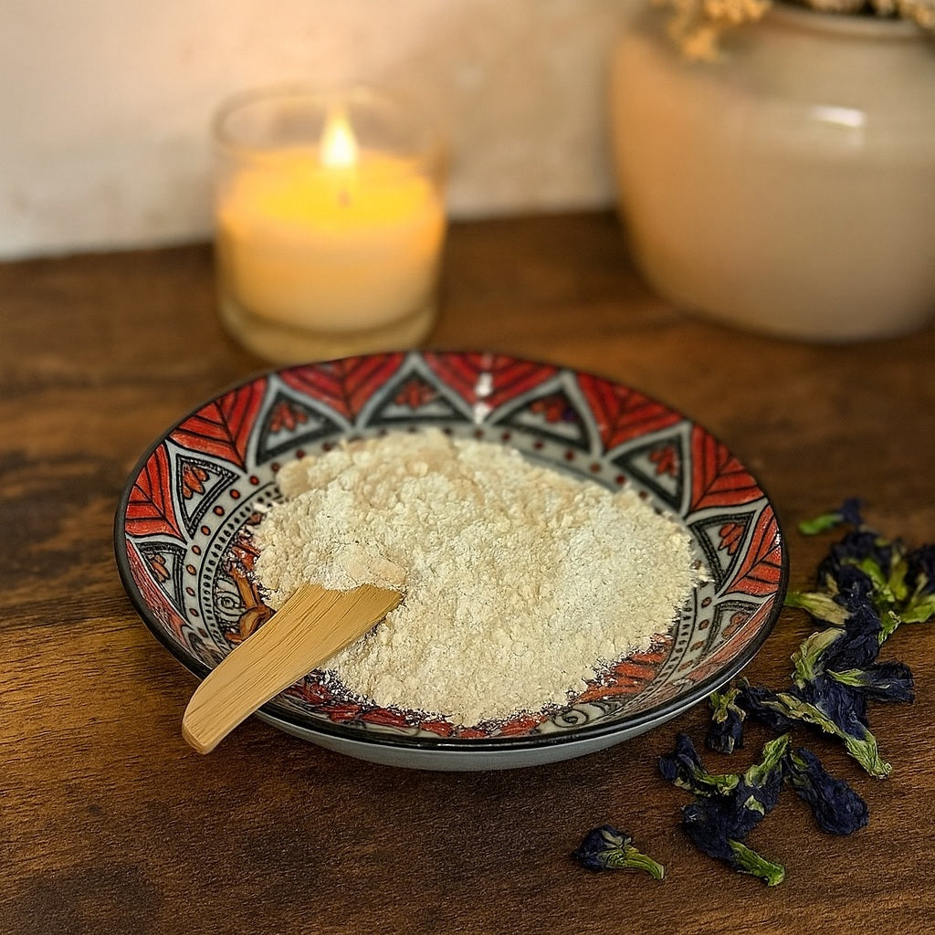 A patterned ceramic plate filled with fine white kaolin clay powder, accented with dried butterfly pea flowers, placed on a rustic wooden vanity with warm, soft lighting and subtle country chic décor in the background.