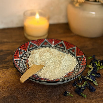 A patterned ceramic plate filled with fine white kaolin clay powder, accented with dried butterfly pea flowers, placed on a rustic wooden vanity with warm, soft lighting and subtle country chic décor in the background.