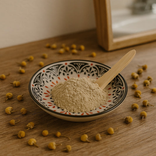 Ceramic bowl with pale yellow French clay and wooden spatula, surrounded by dried chamomile flowers on a rustic wooden bathroom counter.